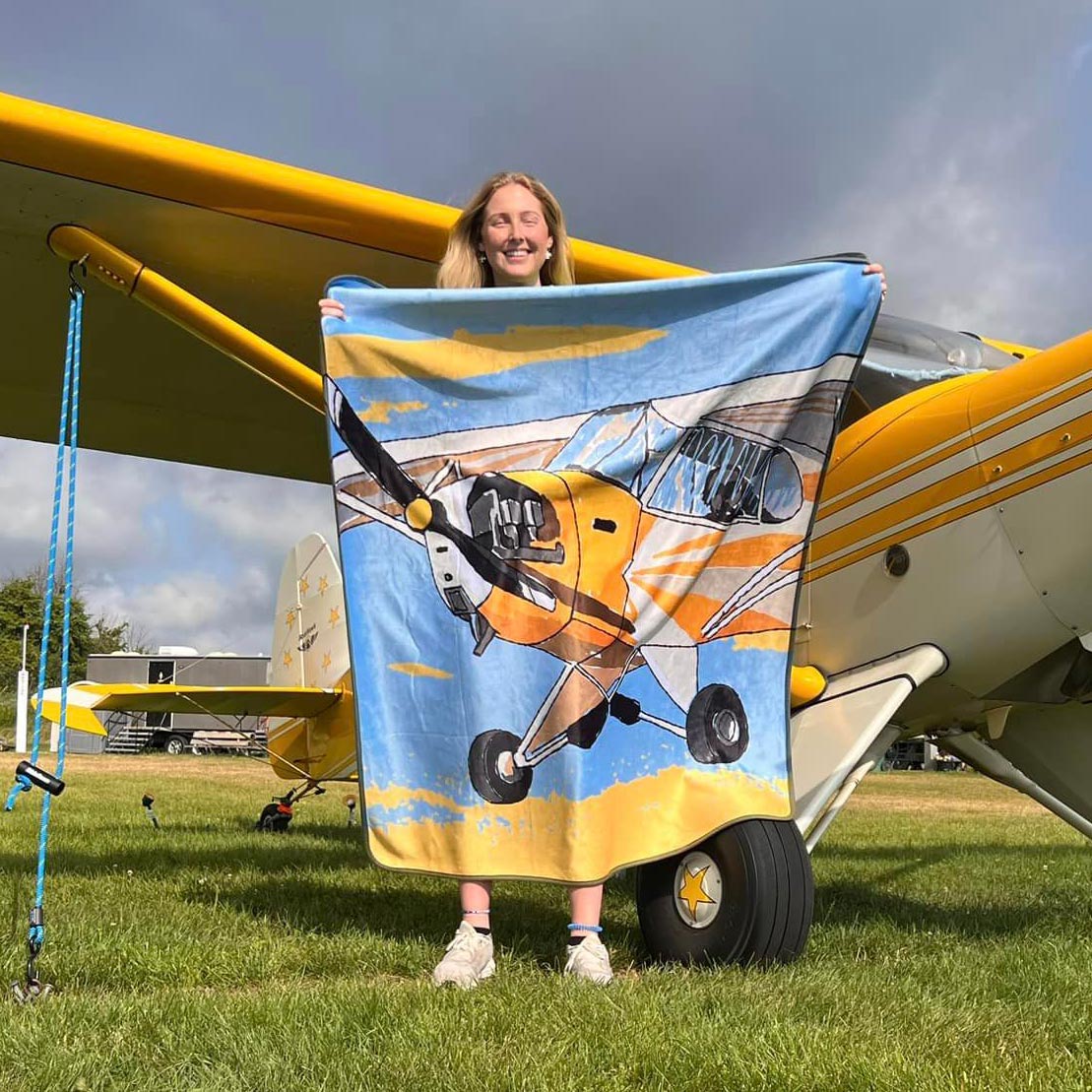 Person holding a yellow Piper J-3 Cub custom fleece blanket beside a matching yellow J-3 Cub airplane, highlighting aviation-themed textile artwork and personalized pilot gift design.