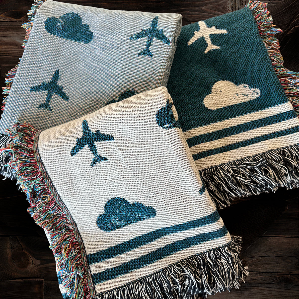 Three woven travel blankets with airplane and cloud patterns with blue and gray tones.