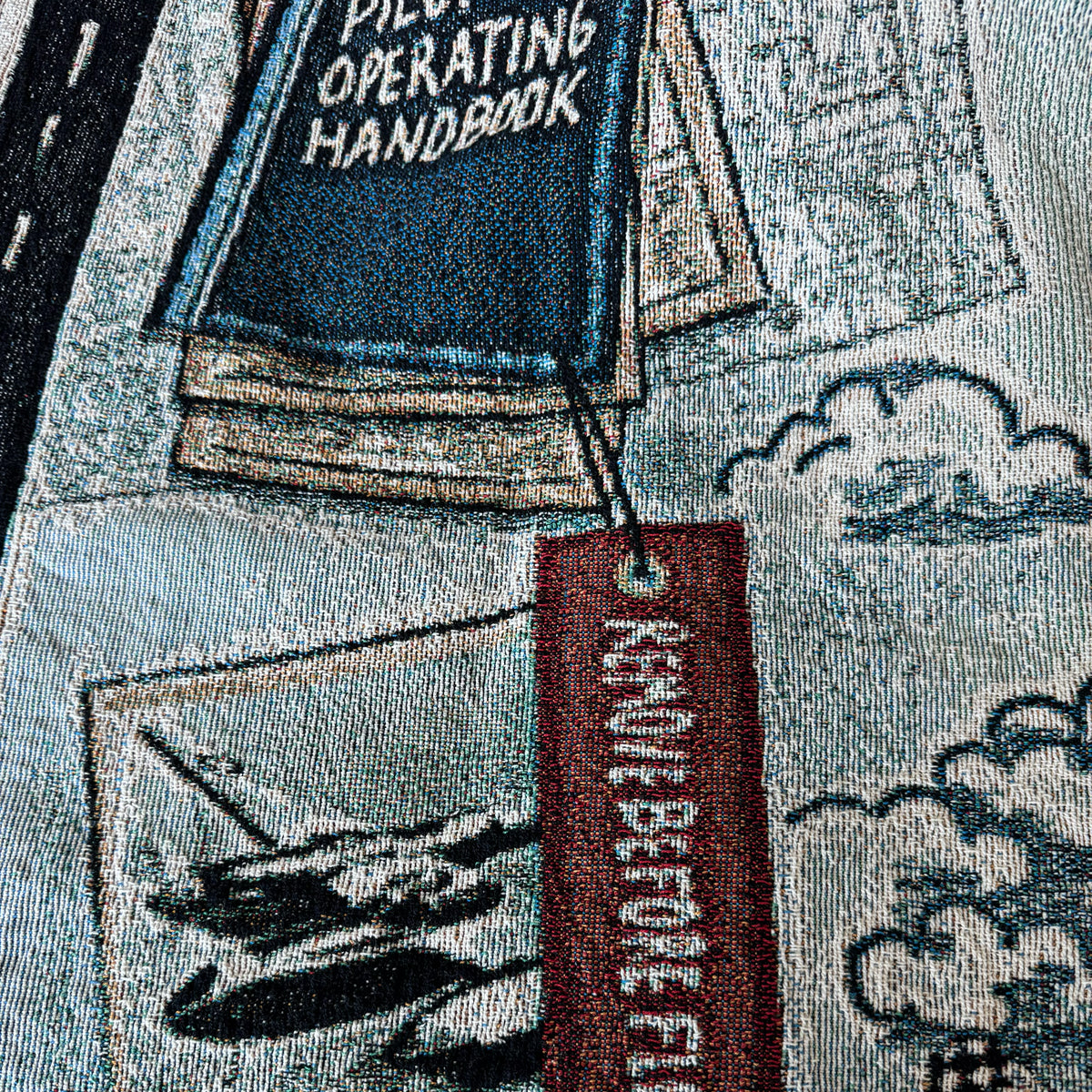 Woven blanket closeup with 'Pilot Operating Handbook' tag and "remove before flight" tag airplane illustration