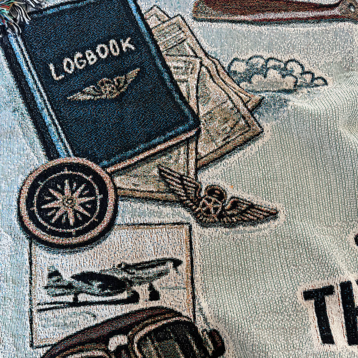 Closeup of woven blanket with aviation-themed design including a logbook, airplane, and P-51 Mustang on a fabric background.