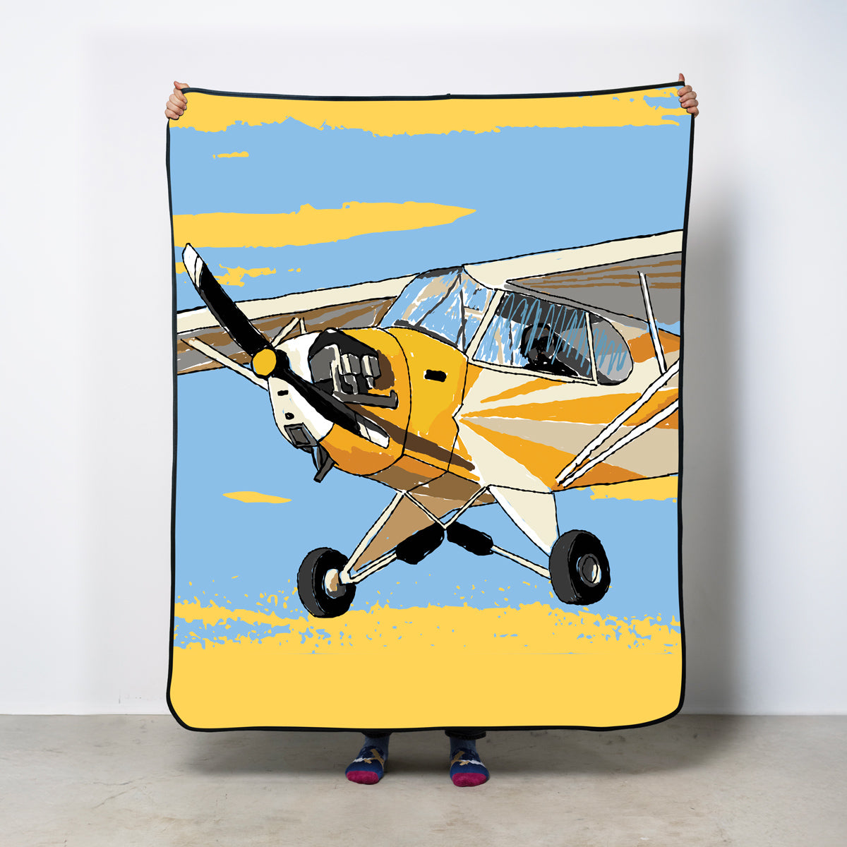 Person holding a blanket with a yellow airplane design against a sky background