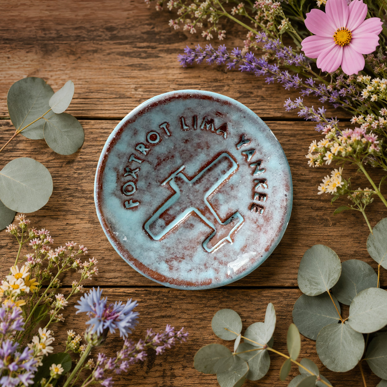 Decorative ceramic trinket dish with 'Foxtrot Lima Yankee' engraving on a wooden surface with flowers and leaves.